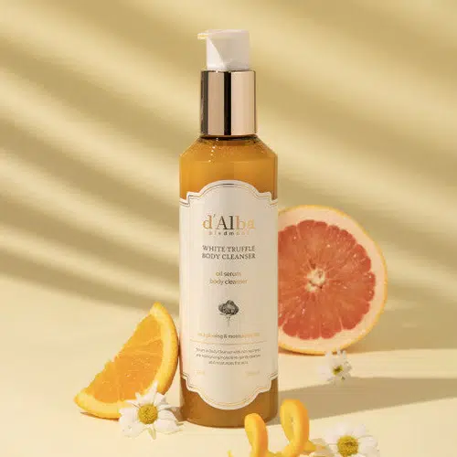 Le flacon de « D'Alba Nettoyant Corps Luxe à la Truffe Blanche et Huile Sérum 275 ml » est représenté debout, entouré de tranches de pamplemousse et d'orange ainsi que de fleurs de camomille, sur un fond jaune pâle parsemé d'ombres délicates. Dakar