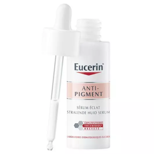 Un flacon blanc de Sérum éclat anti-pigments Eucerin avec un bouchon compte-gouttes, avec des accents roses et gris et un texte soulignant son ingrédient breveté Thiamidol. Le compte-gouttes repose à côté du flacon ouvert. Dakar