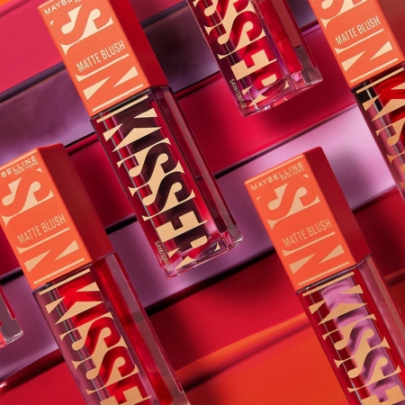 Plusieurs flacons de « Sunkisser Matte Blush Liquide Effet Mat » de Maybelline New York sont disposés en diagonale sur un fond rose vif et orange, chacun arborant un texte graphique et audacieux sur son emballage. Dakar