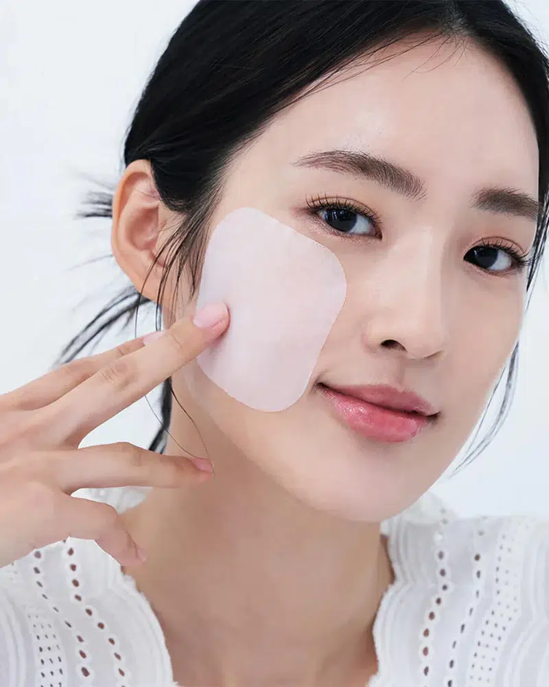 Une femme à la peau claire, vêtue d'un haut blanc texturé, sourit doucement à la caméra alors qu'elle applique un patch Mediheal Disques de Soin Masque Éclaircissants et Purifiants 100 Pièces sur sa joue. Dakar