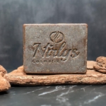 Une barre rectangulaire Nirlys Paris Savon 2 en 1 Exfoliant et Nettoyant 200 g avec la marque et le logo gravés repose sur une surface texturée en pierre brune, entourée de pierres similaires, soulignant son attrait luxueux. Dakar