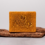 Une barre de savon rectangulaire orange-brun avec "Nolys" et un logo gravé est présentée sur du bois texturé, probablement le Nirlys Paris Savon Illuminatrice et Anti Taches Au Curcuma Carotte à la Vitamine C 200 g. Dakar
