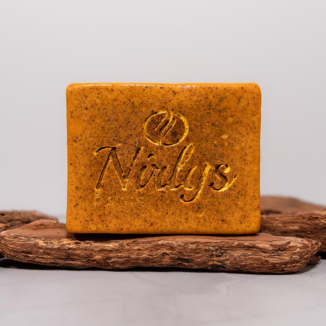 Une barre de savon rectangulaire orange-brun avec "Nolys" et un logo gravé est présentée sur du bois texturé, probablement le Nirlys Paris Savon Illuminatrice et Anti Taches Au Curcuma Carotte à la Vitamine C 200 g. Dakar