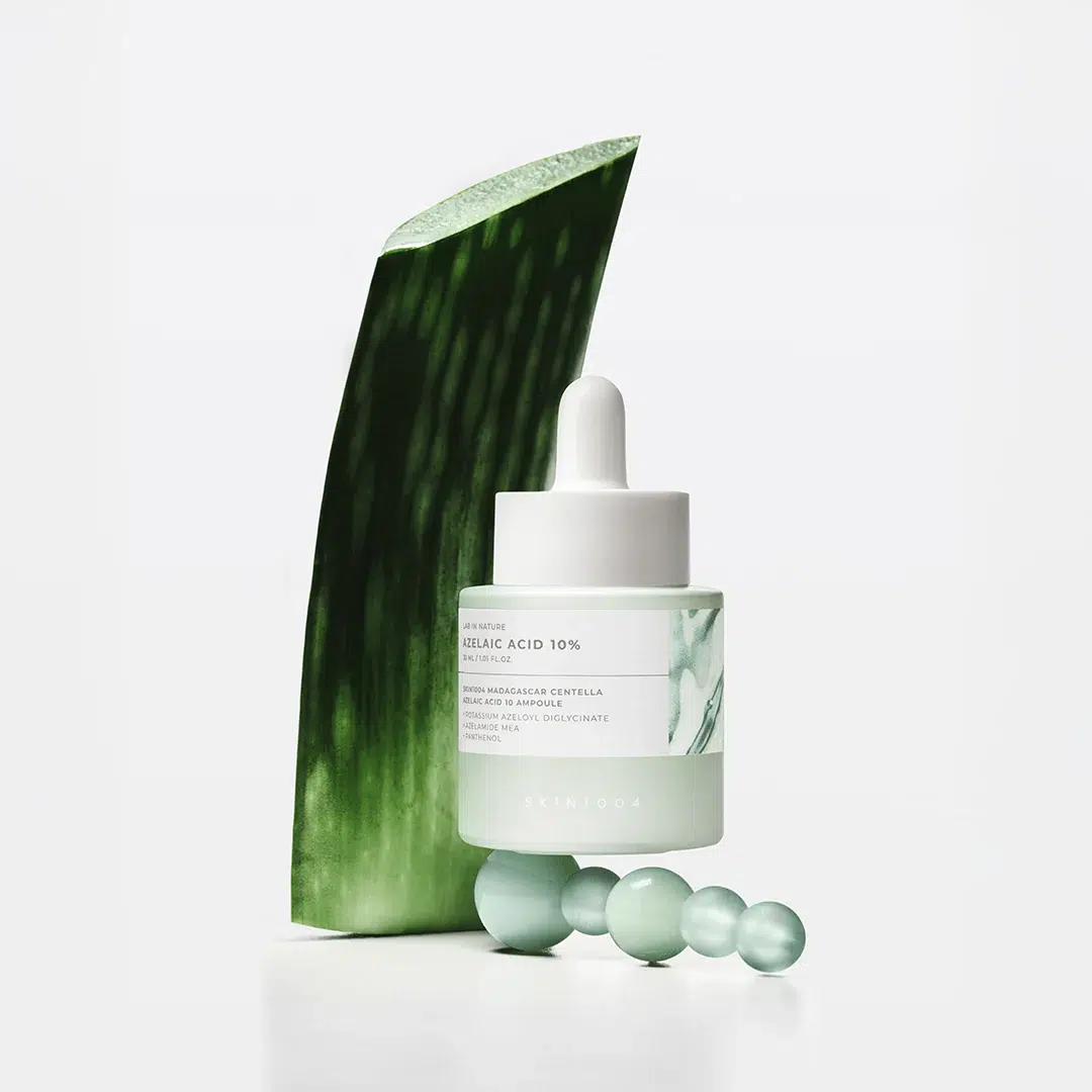Un flacon compte-gouttes de Skin1004 « Azelaic Acid Ampoule Soin Éclat Anti-Rougeurs et Anti-Taches » de 30 ml, orné d'une étiquette blanche et verte, est placé devant une grande feuille d'aloe vera, entouré de quatre perles de verre vertes sur un fond blanc immaculé. Dakar