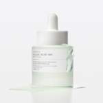 Un flacon compte-gouttes blanc contenant l'ampoule « Skin1004 Azelaic Acid Ampoule Soin Éclat Anti-Rougeurs et Anti-Taches » de 30 ml repose sur une surface blanche, entouré d'un liquide vert clair. Son emballage minimaliste présente un motif inspiré du marbre. Dakar