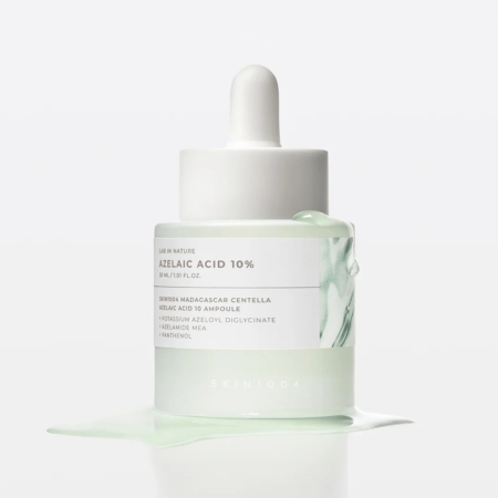 Un flacon compte-gouttes blanc contenant l'ampoule « Skin1004 Azelaic Acid Ampoule Soin Éclat Anti-Rougeurs et Anti-Taches » de 30 ml repose sur une surface blanche, entouré d'un liquide vert clair. Son emballage minimaliste présente un motif inspiré du marbre. Dakar