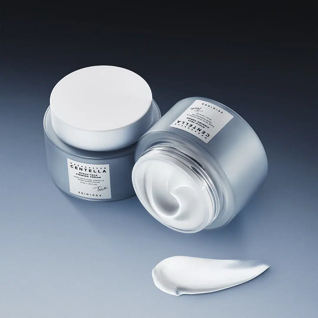 Deux pots de crème « Hyalu Teca Crème Anti-Rides et Peptides Peau Plus Ferme » de Skin1004, de 50 ml chacun — l’un fermé, l’autre ouvert, laissant apparaître une crème blanche et onctueuse —, avec une noisette de produit à côté. Les pots argentés et blancs arborent des étiquettes minimalistes. Dakar