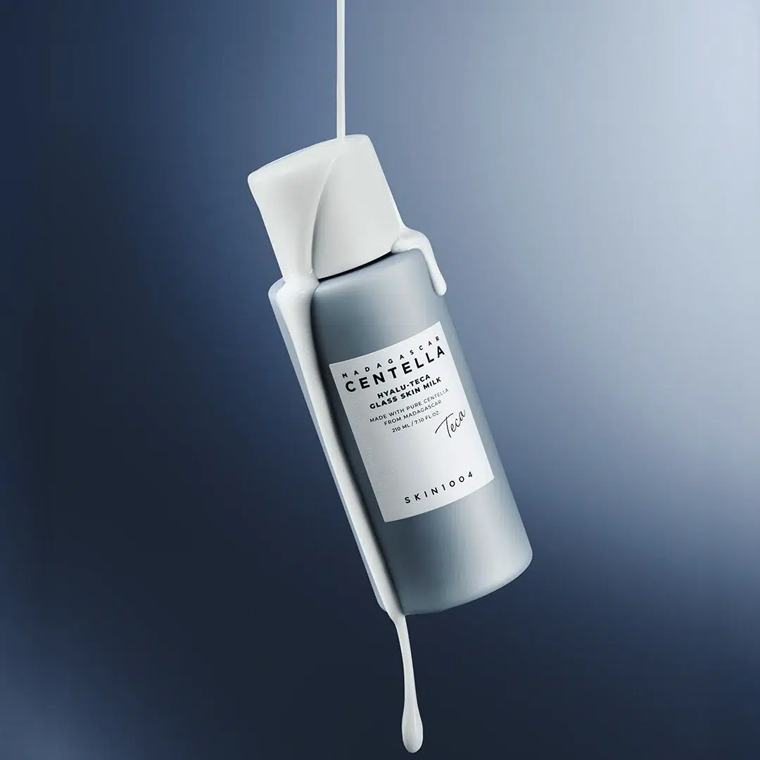 Un flacon de Skin1004 Hyalu Teca Glass Skin Milk Essence Lactée Hydratante et Éclat de 210 ml flotte dans les airs, tandis qu’un liquide blanc ruisselle sur un fond bleu dégradé. Dakar