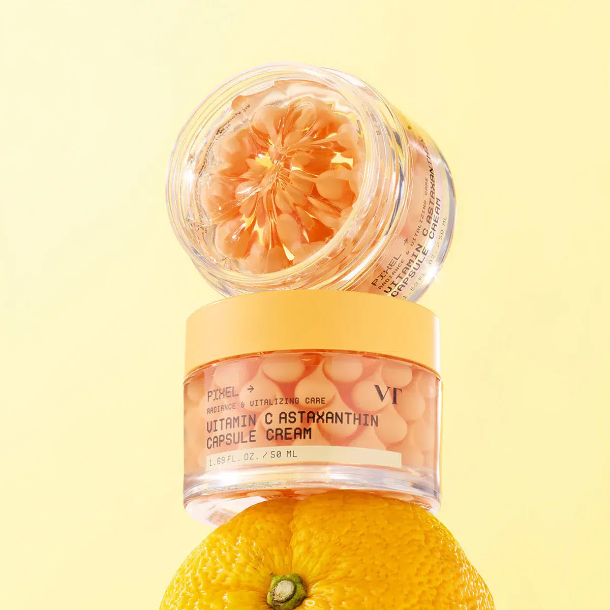 Deux pots de Vt Crème Éclat aux capsules de vitamine C et d'astaxanthine, anti-taches et anti-âge (50 ml), sont empilés sur une orange, sur un fond jaune clair. Le pot du haut est ouvert, laissant apparaître ses capsules d'un orange vif. Dakar