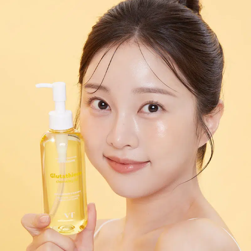 Une femme à la peau éclatante et lumineuse sourit doucement tout en présentant l'huile démaquillante Vt Éclat et Hydratation au Glutathion 200 ml sur un fond jaune uni. Dakar