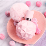 Un flacon rose à facettes de l'eau de parfum pour femme « Sweet Like Candy » d'Ariana Grande, d'une contenance de 100 ml, orné d'un pompon duveteux, repose sur un plateau rose en forme de cœur, entouré de bonbons roses et blancs posés sur une surface en marbre. Dakar