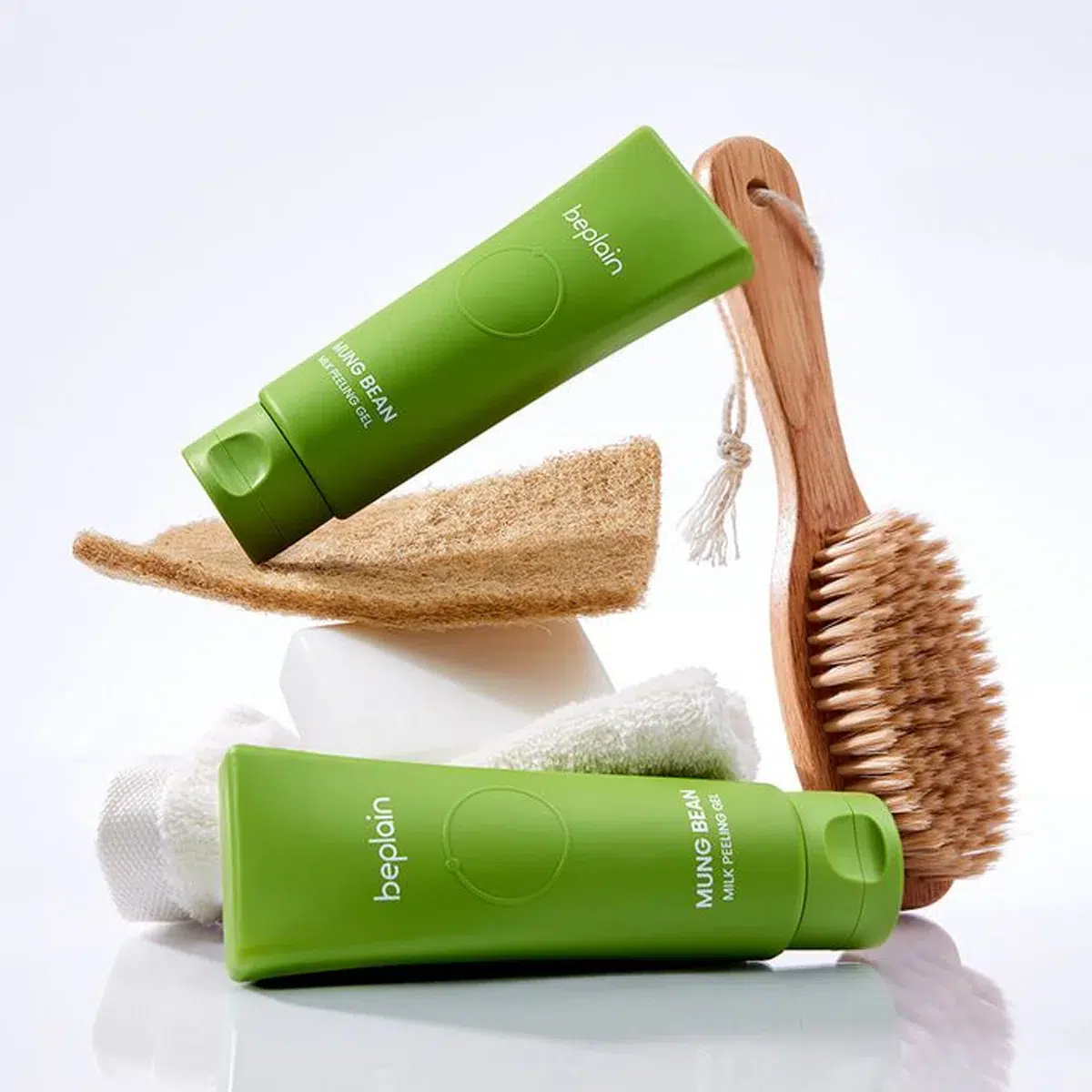 Deux tubes verts de « Beplain Gel Exfoliant au Lait de Haricot Mungo Peaux Sensibles » de 120 ml, une brosse en bois, un éponge en loofah, une serviette blanche et un savon en pain sont soigneusement disposés sur une surface blanche devant un fond clair. Dakar