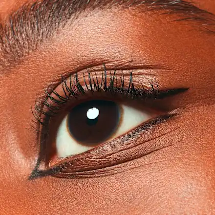 Gros plan sur l'œil brun d'une personne aux cils longs et volumineux, mis en valeur grâce au mascara Essence Lash Without Limits Waterproof 03 Black Allongeant et Volumisant 13 ml, qui fait ressortir la douceur de la peau mate et un sourcil bien dessiné, souligné par un trait d'eyeliner noir intense. Dakar