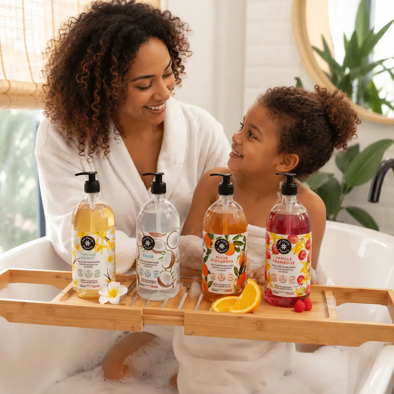 Une femme et un enfant, dans un bain moussant, se sourient. Quatre flacons colorés de savon ou de gel douche Univers Cosmetix sont disposés sur un plateau de bain en bambou, accompagnés de tranches de fruits et de fleurs. Dakar