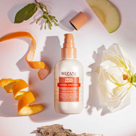 Un flacon de crème lissante thermique « Press Agent » de Mizani est entouré d'un zeste d'orange, d'une rose blanche, d'une tranche de pomme verte et de feuilles vertes, le tout disposé sur un fond clair aux ombres douces. Dakar