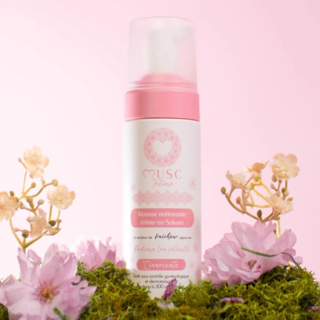 Un flacon de « Musc Intime Fleur de Sakura L’innocente Mousse Nettoyante Intime » de 150 ml est présenté au milieu de fleurs roses et de mousse, sur un fond rose pâle. L’emballage se décline dans des tons blancs et roses, agrémenté d’élégants motifs floraux. Dakar