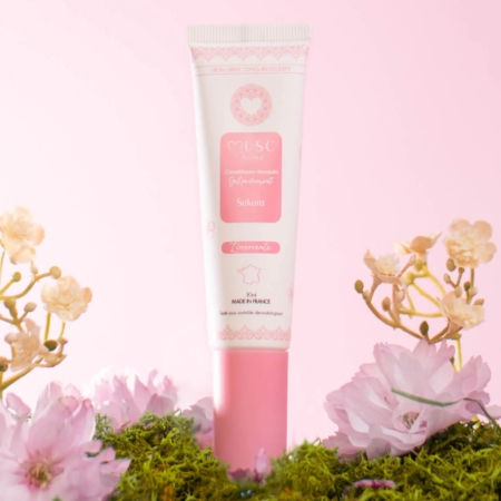 Un tube de cosmétique rose et blanc portant l'inscription « Sakura » se dresse au milieu de la mousse et des fleurs roses, sur un fond rose pâle. Dakar