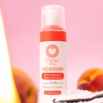 Un flacon de 150 ml de « Musc Intime Mousse Nettoyante Intime Hot Peach La charmeuse » trône sur une surface rose, entouré de pêches mûres et d'une gousse de vanille en arrière-plan. Le flacon blanc, rehaussé de touches couleur pêche et d'un logo en forme de cœur, met en valeur son charme délicat. Dakar