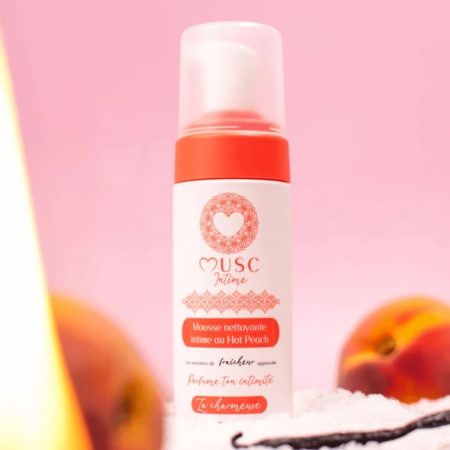 Un flacon de 150 ml de « Musc Intime Mousse Nettoyante Intime Hot Peach La charmeuse » trône sur une surface rose, entouré de pêches mûres et d'une gousse de vanille en arrière-plan. Le flacon blanc, rehaussé de touches couleur pêche et d'un logo en forme de cœur, met en valeur son charme délicat. Dakar