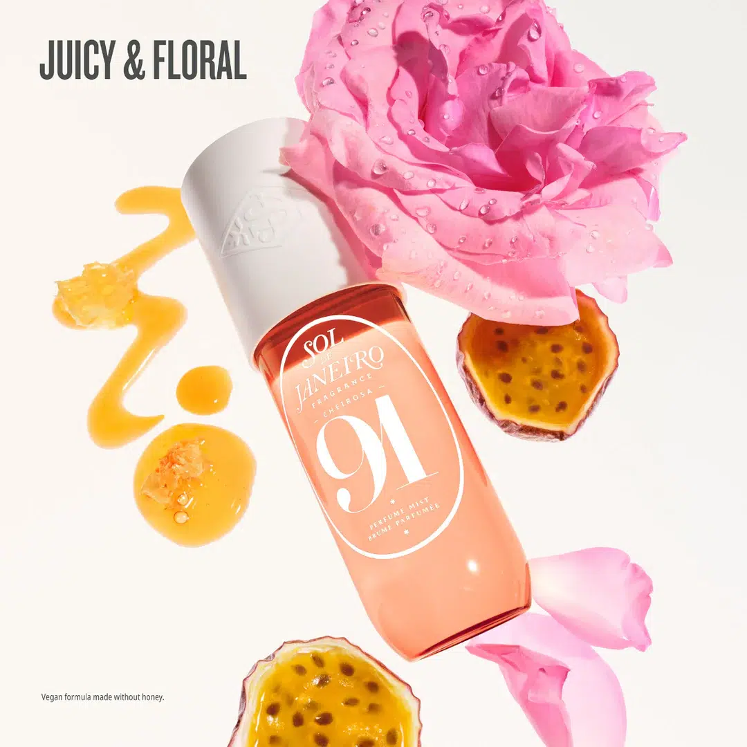 Un flacon de Sol de Janeiro Cheirosa 91 Brume Parfumée Corps et Cheveux de 90 ml repose à côté d’une rose rose, de moitiés de fruit de la passion et d’une goutte de liquide jaune. La mention « JUICY & FLORAL » est mise en avant, tandis qu’une note en petits caractères précise que la formule est végane et ne contient pas de miel. Dakar