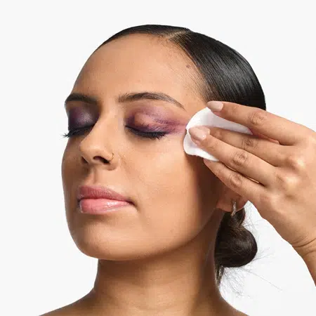 Une femme aux cheveux foncés tirés en arrière, l'air serein, démaquille délicatement son fard à paupières violet à l'aide d'un coton et du démaquillant Clinique Take The Day Off pour les yeux, les cils et les lèvres, sans parfum, 125 ml. Elle a les yeux fermés tandis qu'elle se démaquille. Dakar